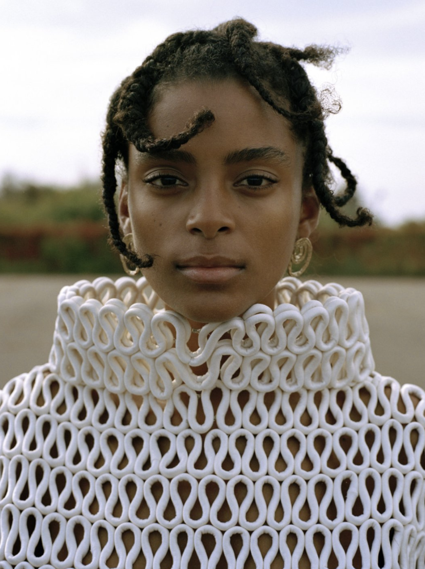 Close-up portrait of a young woman with braided hair, wearing a white sculptural top, outdoors under a cloudy sky. Fashion photography.
