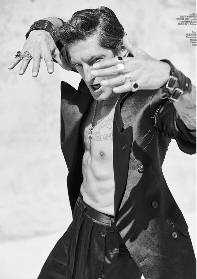 Portrait of a male fashion model with dark hair, wearing a black suit jacket and trousers, accessorized with jewelry, striking a pose.