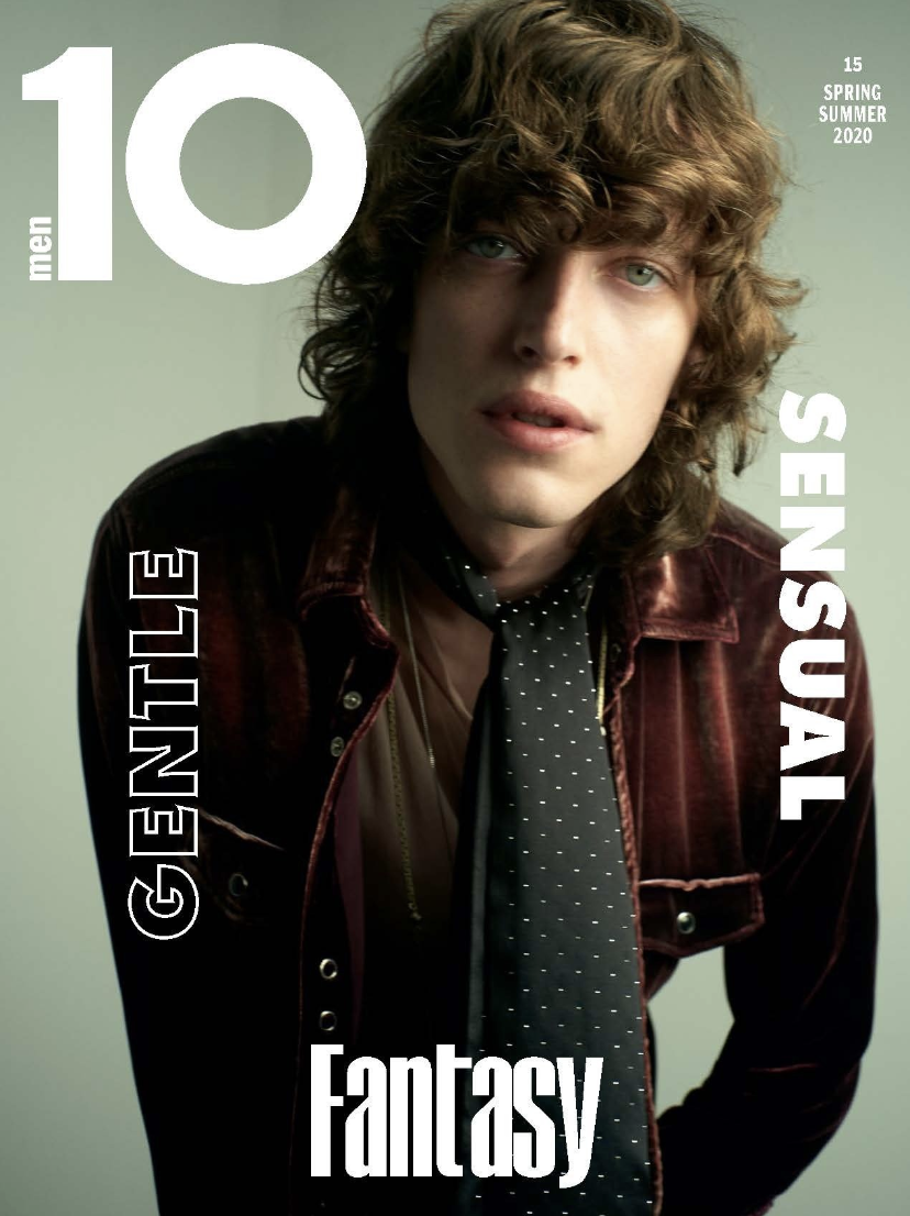 Magazine cover featuring a fair-skinned male model with shoulder-length wavy brown hair, wearing a velvet jacket and patterned tie.