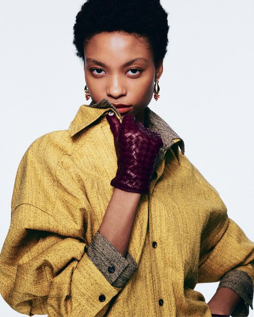 Close-up portrait of a dark-skinned woman in a mustard-colored oversized shirt and leather gloves, posing in a studio setting.