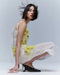 Model in white dress with yellow textured embellishments and black heels crouched against white background