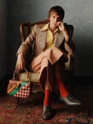 Young man seated in a vintage armchair holding a color-blocked handbag, styled by Jordan Boorman.