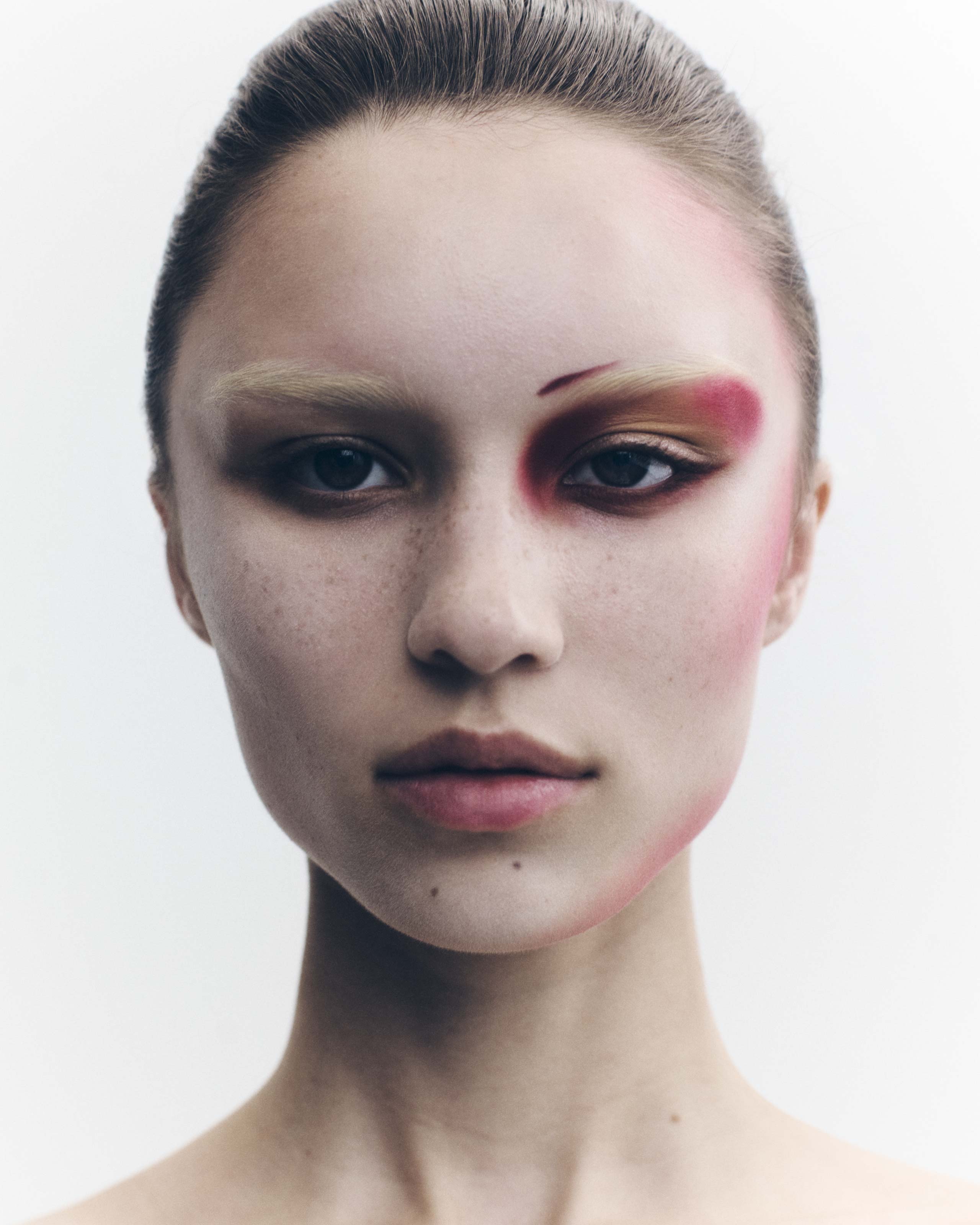 Close-up portrait of a young woman with unique makeup, including red and brown eyeshadow and blush, against a bright white backdrop.