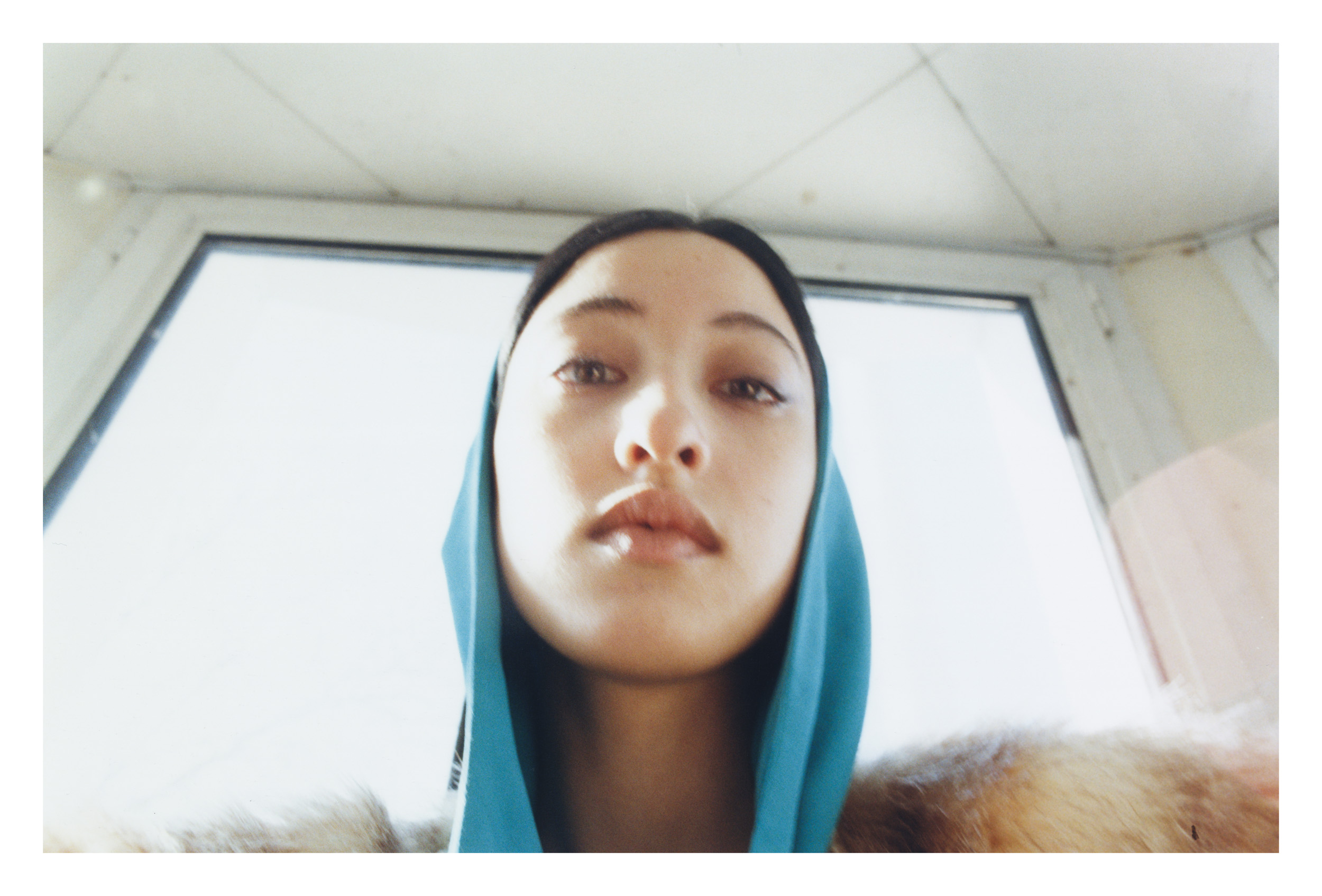 Close-up portrait of an adult female with dark hair, wearing a blue headscarf and fur coat, looking directly at the camera in an indoor setting.
