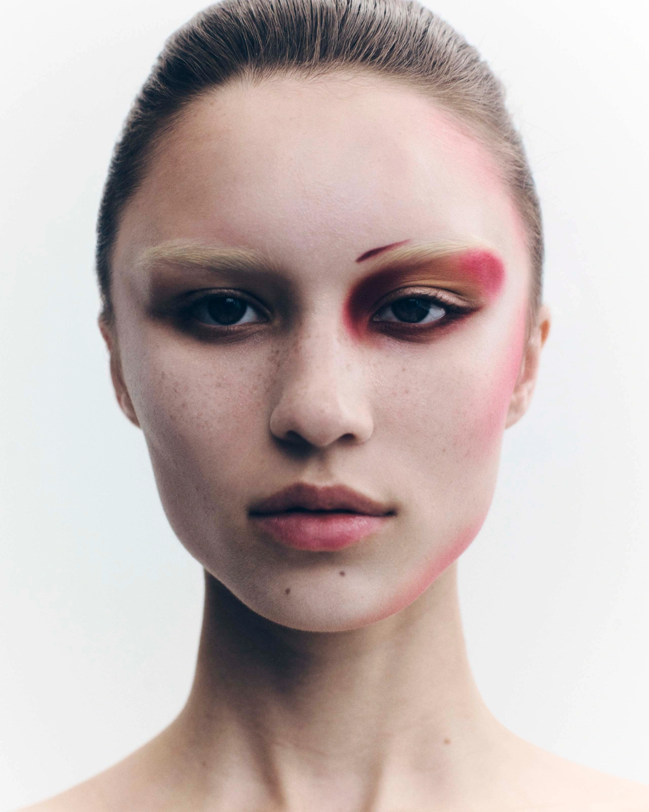 Close-up portrait of a woman with bleached eyebrows and striking red and black editorial makeup against a white backdrop.