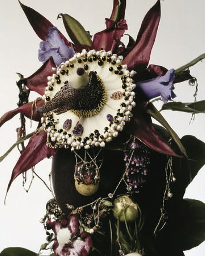 Floral arrangement featuring an array of flowers in purples, greens, and reds along with jewelry details and decorative elements.