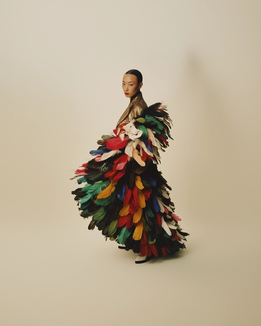 Asian woman in gold jacket posing, adorned with colorful feathered dress. Neutral background, fashion editorial photography.
