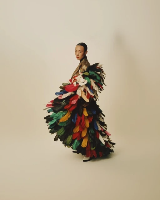Asian woman in gold jacket posing, adorned with colorful feathered dress. Neutral background, fashion editorial photography.