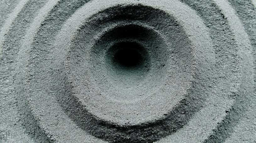 Concentric circles carved into gray sand, forming a textured pattern with a dark central void, shot in natural light.