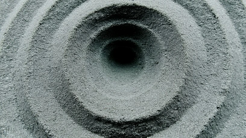 Concentric circles carved into gray sand, forming a textured pattern with a dark central void, shot in natural light.