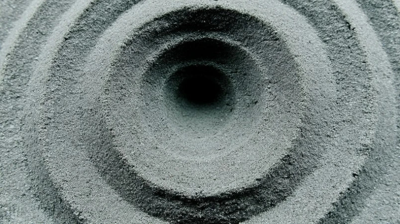 Concentric circles carved into gray sand, forming a textured pattern with a dark central void, shot in natural light.