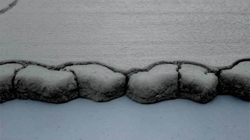 Close-up of a cracked, weathered concrete surface with an irregular textured edge, creating an abstract, monochromatic landscape.