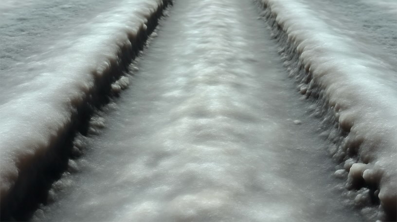Close-up of a textured, light-grey and white salt flat with linear tire tracks running through it, shot with a low perspective.