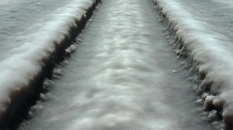 Close-up of a textured, light-grey and white salt flat with linear tire tracks running through it, shot with a low perspective.