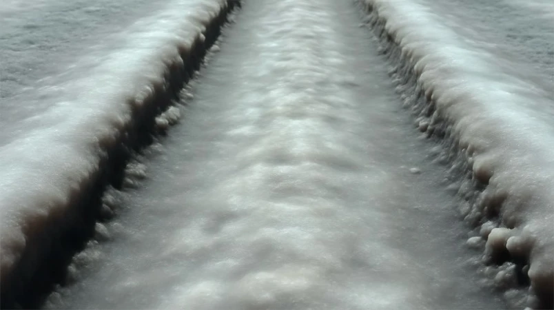 Close-up of a textured, light-grey and white salt flat with linear tire tracks running through it, shot with a low perspective.