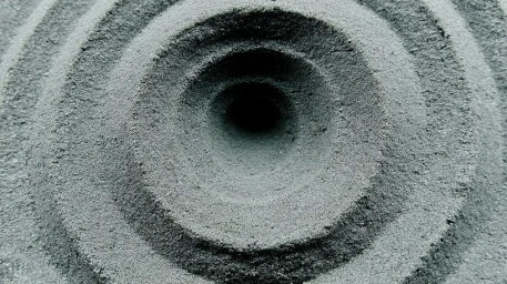 Close-up of concentric circular ripples carved into textured grey sand creating a deep central vortex.