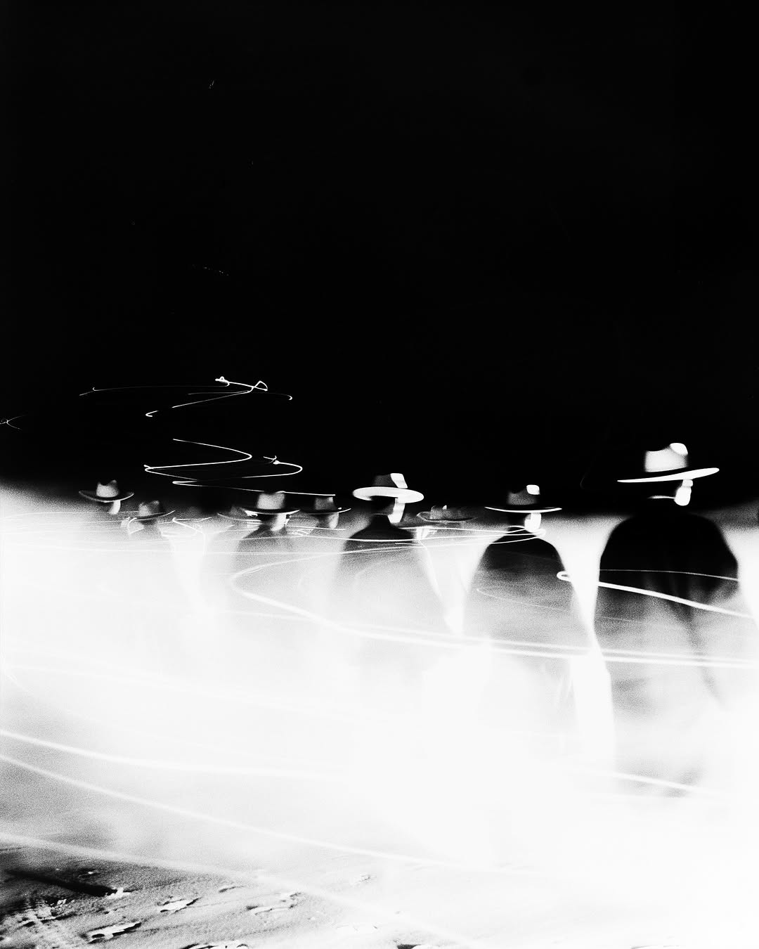 Monochrome long exposure of a group of adult males walking in hats with motion blur and light trails on a dark background.