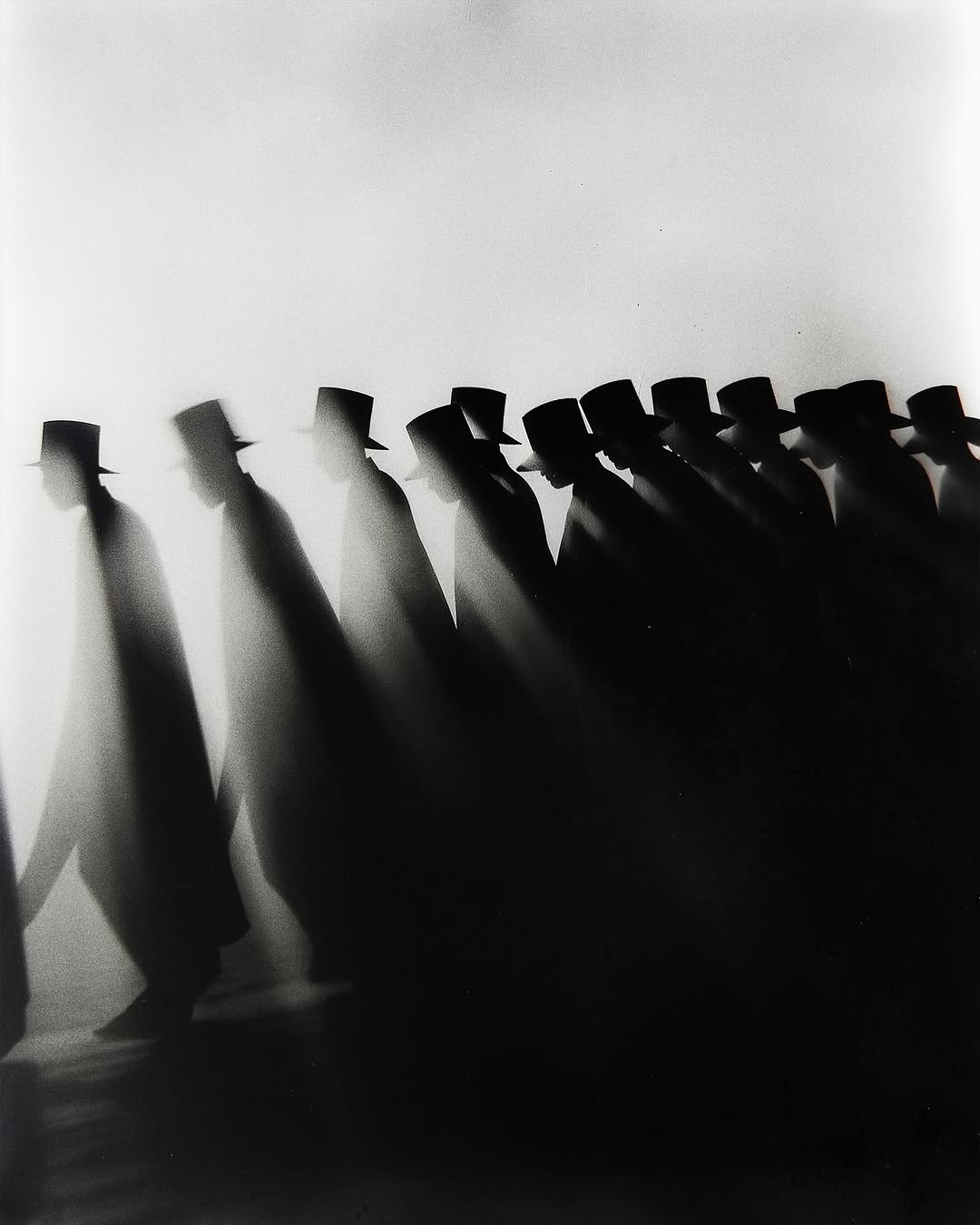 Black and white photo of a long line of silhouetted figures wearing hats, walking in the same direction against a bright background.