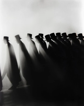 Black and white photo of a long line of silhouetted figures wearing hats, walking in the same direction against a bright background.