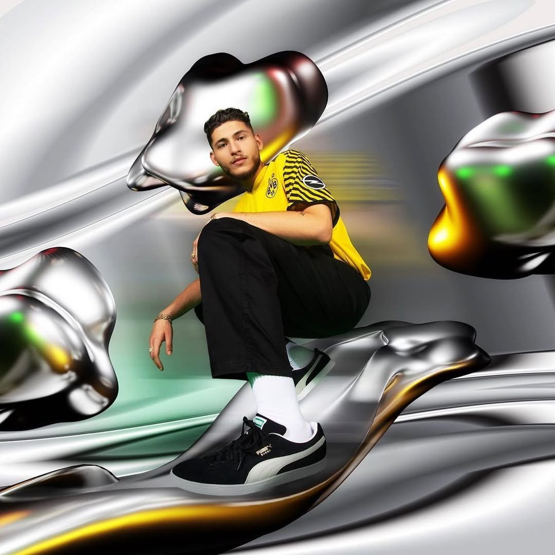 Young man in yellow and black soccer jersey poses with black Puma sneakers against a metallic liquid background, glossy textures.