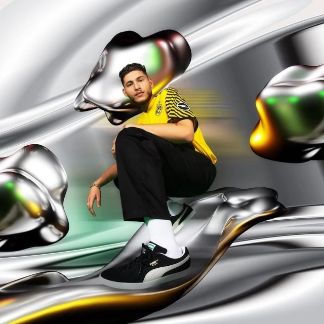 Young man in yellow and black soccer jersey poses with black Puma sneakers against a metallic liquid background, glossy textures.