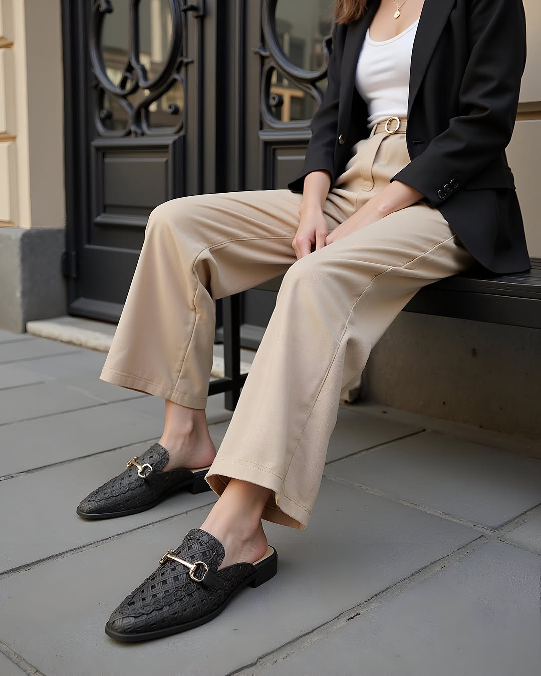 Woman in black blazer and beige pants sits wearing woven black mules with gold hardware detail in an outdoor, urban setting.
