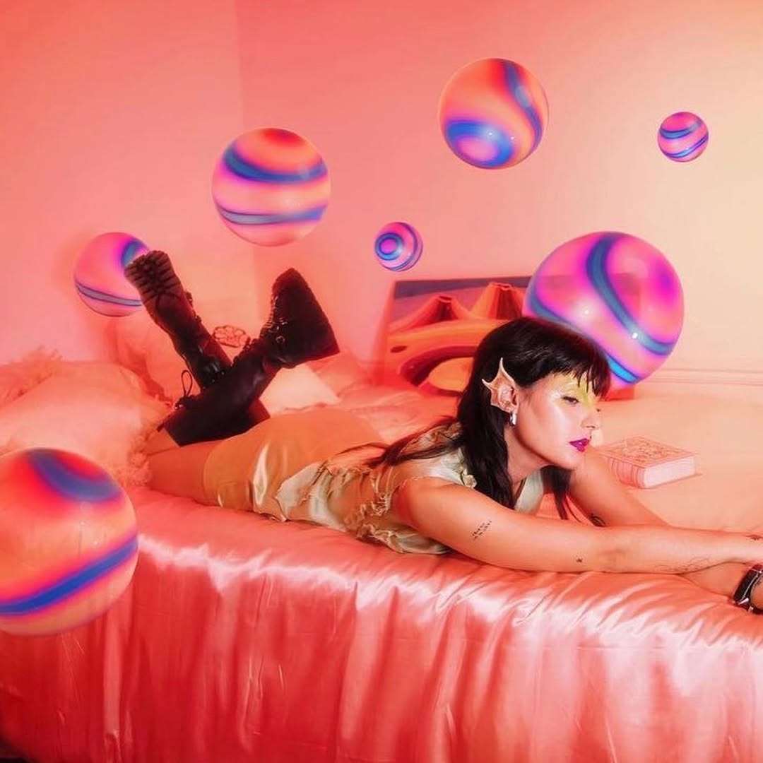 Woman in fantasy-inspired attire poses on pink bed amid floating, striped spheres and fairylike accessories in brightly lit indoor photo.