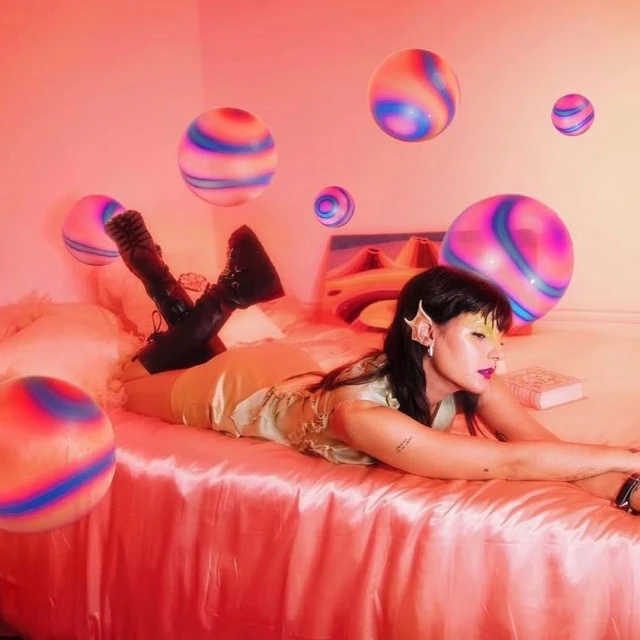 Woman in fantasy-inspired attire poses on pink bed amid floating, striped spheres and fairylike accessories in brightly lit indoor photo.