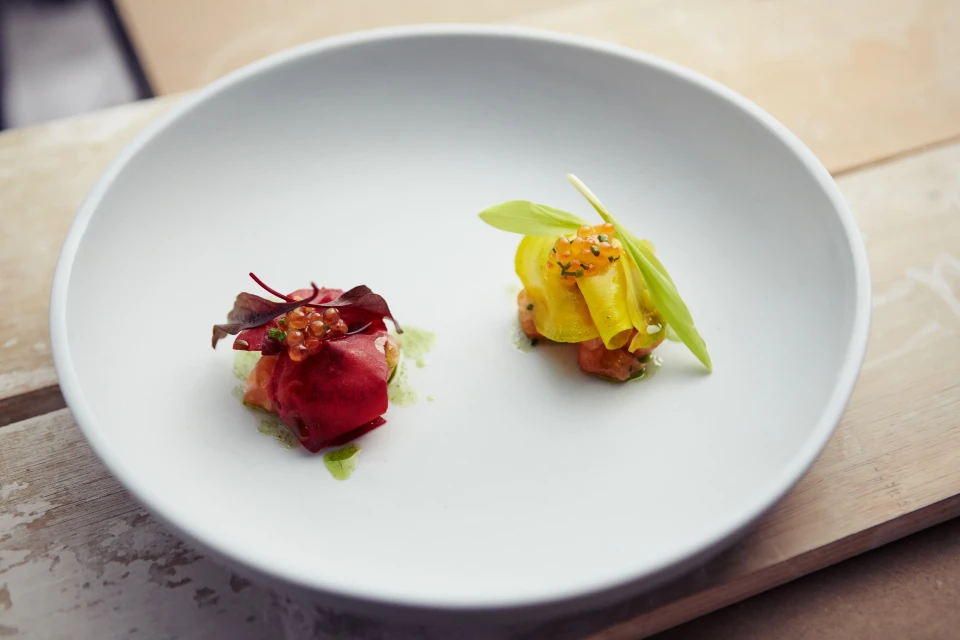 Two petite culinary artworks featuring vibrant red and yellow vegetable compositions artfully arranged on a pristine white plate, on a wood surface.