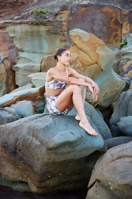 Woman in a brown and white patterned two-piece outfit sitting on a large rock formation near the beach, posing in natural light.