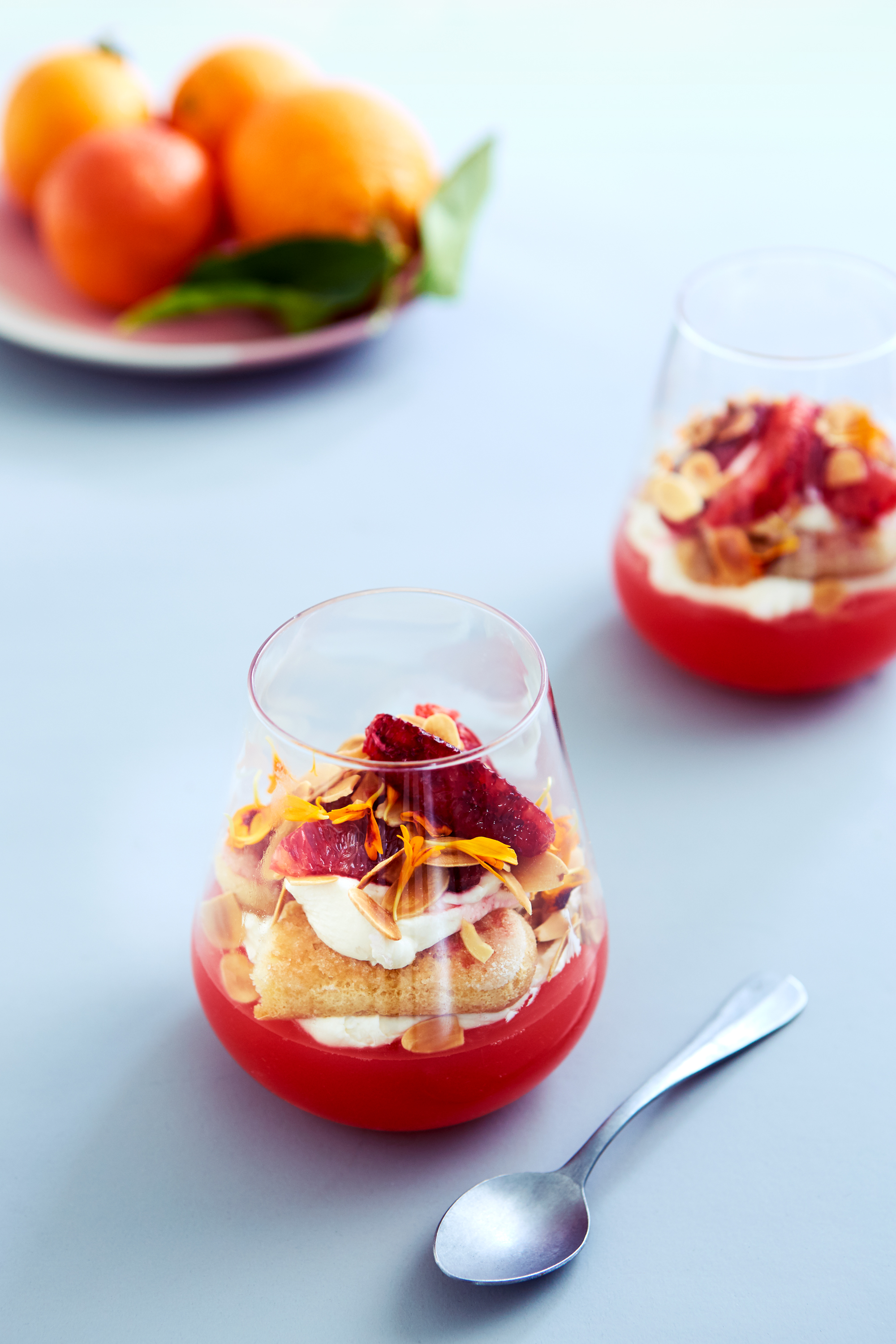 A glass dessert with layers of red jelly, cream, cake, almonds, and citrus zest sits on a gray surface next to a spoon.