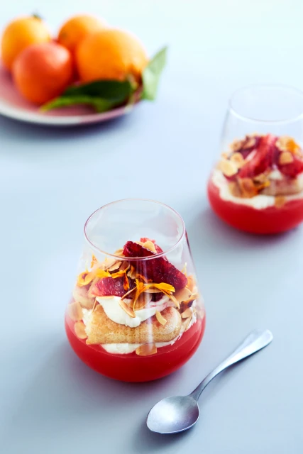 A glass dessert with layers of red jelly, cream, cake, almonds, and citrus zest sits on a gray surface next to a spoon.