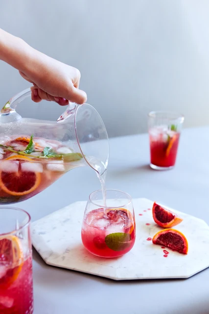 A hand pours blood orange cocktail from a glass pitcher into a tumbler with ice. Marble serving board, bright studio lighting.