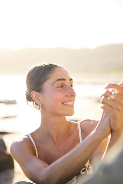Smiling young adult woman with brown hair and a white top on a beach with sunlight in the background, looking to the side.