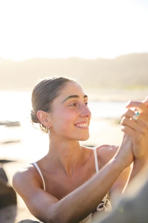 Smiling young adult woman with brown hair and a white top on a beach with sunlight in the background, looking to the side.