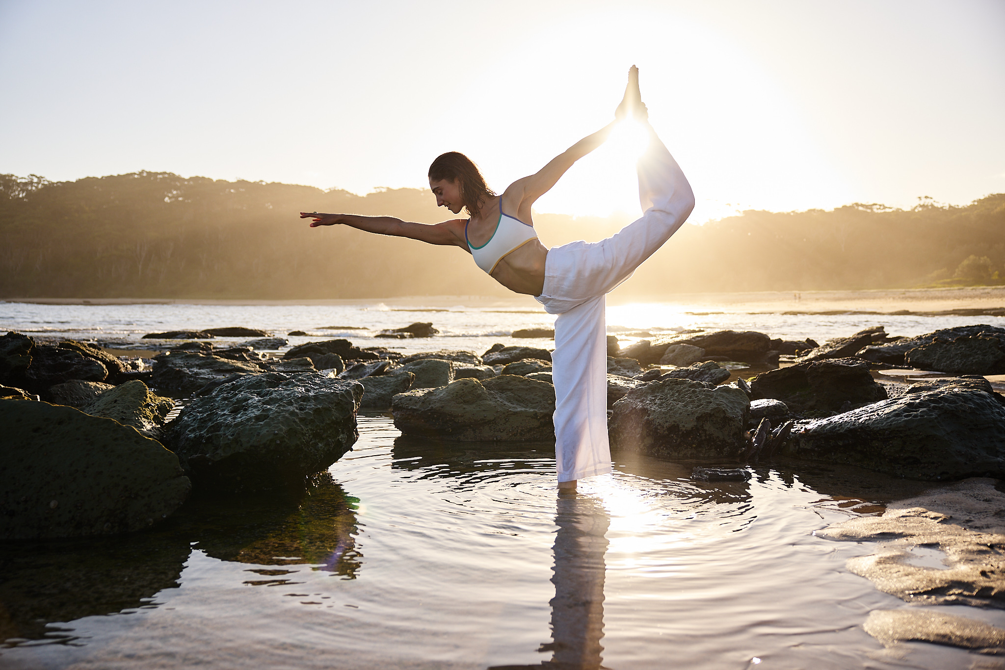 A woman is performing a yoga pose on a rocky beach at sunset with an ocean backdrop. She is wearing a white top and pants.
