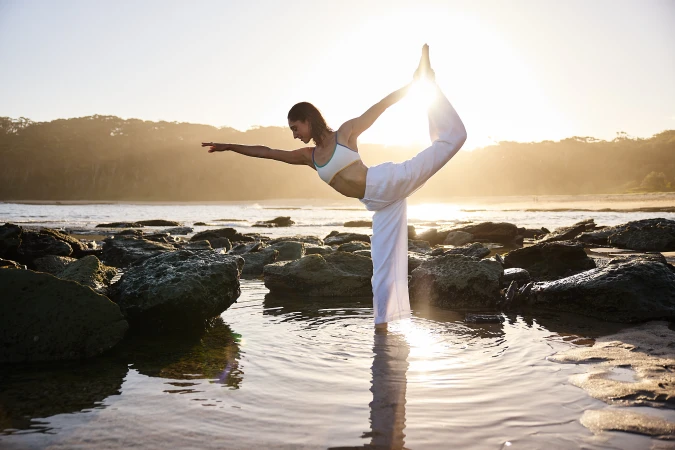 A woman is performing a yoga pose on a rocky beach at sunset with an ocean backdrop. She is wearing a white top and pants.