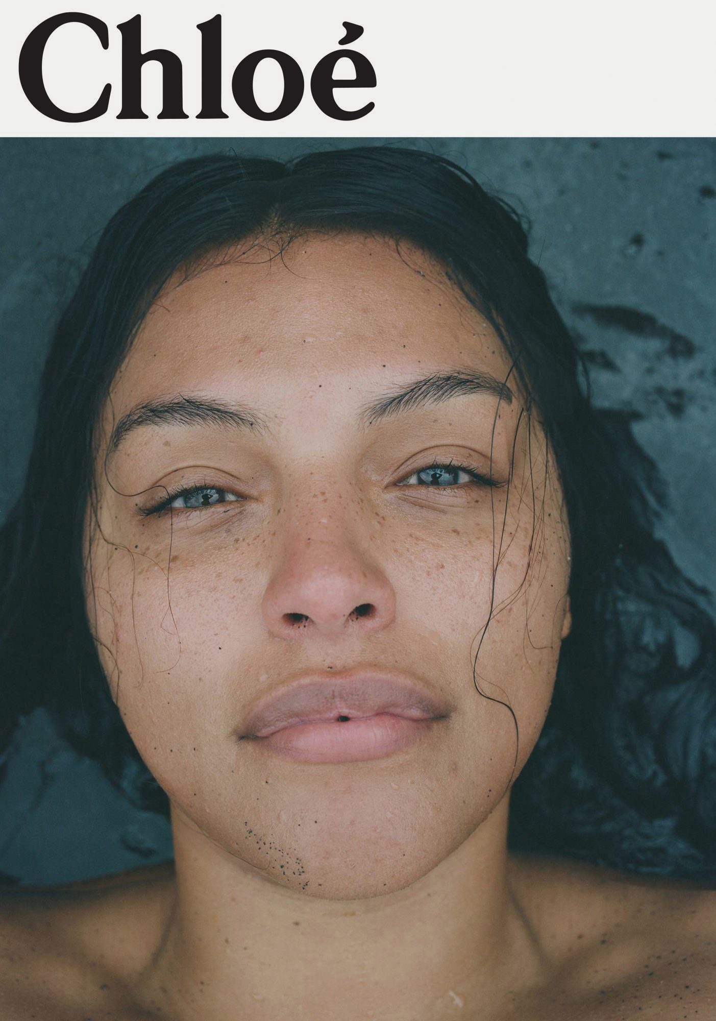 Close-up portrait of a young adult female with wet dark hair, light eyes, and freckles, set against a dark water background. Brand name Chloe visible.
