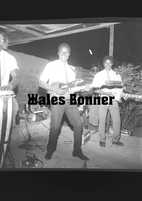 Black and white outdoor shot of three adult musicians performing: one plays drums, two play electric guitars, with Wales Bonner text overlay.