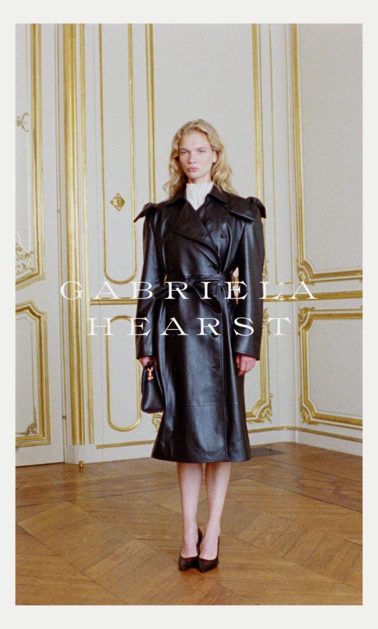 Fashion shot of a blonde woman in a black leather trench coat standing indoors with a purse and heels, posing in front of wall panels.