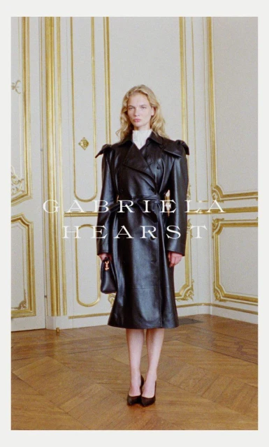 Fashion shot of a blonde woman in a black leather trench coat standing indoors with a purse and heels, posing in front of wall panels.