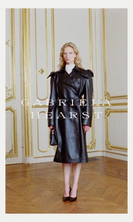 Fashion shot of a blonde woman in a black leather trench coat standing indoors with a purse and heels, posing in front of wall panels.