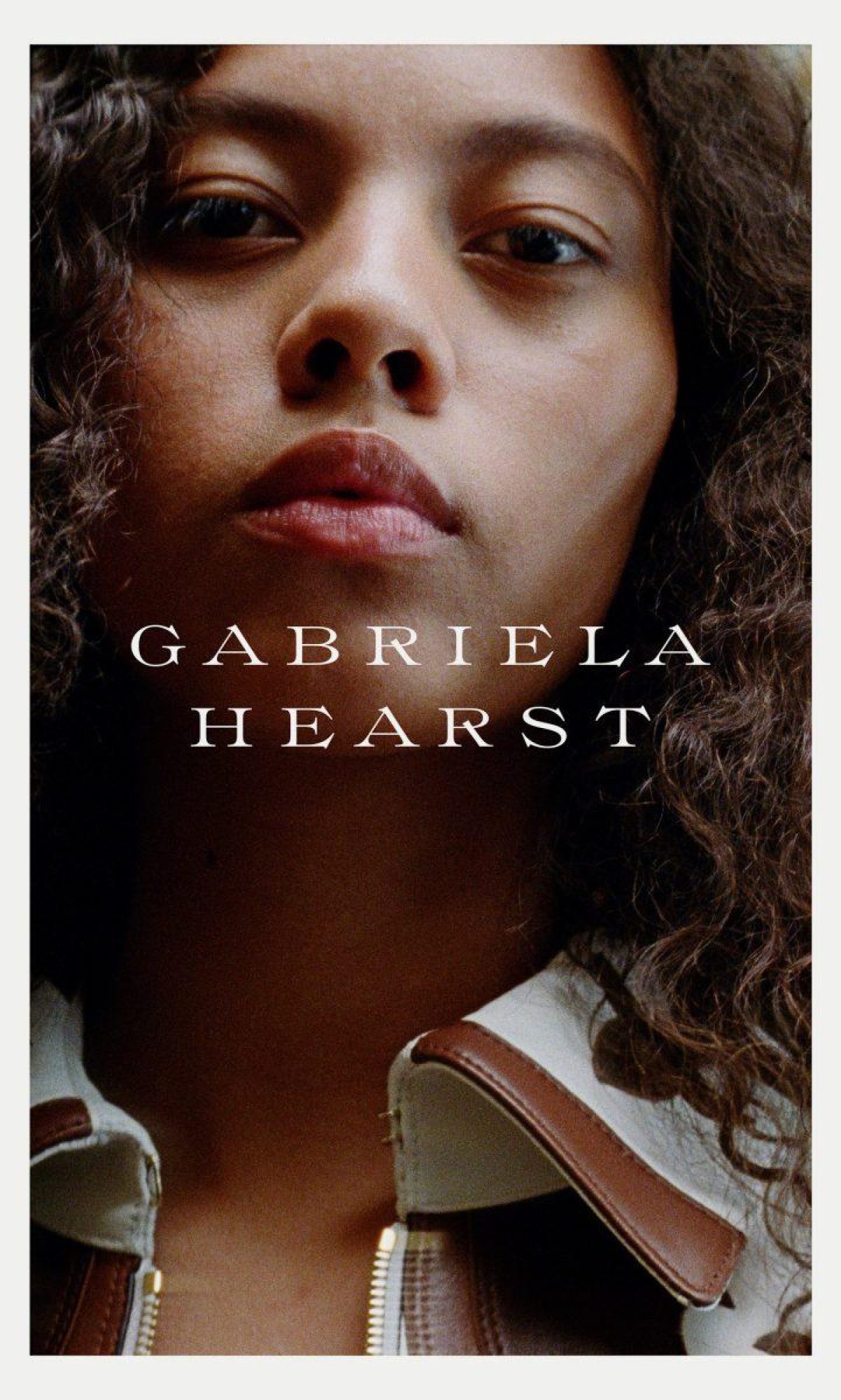 Portrait of a woman with dark curly hair wearing a brown and white jacket, featuring the text GABRIELA HEARST in all caps.