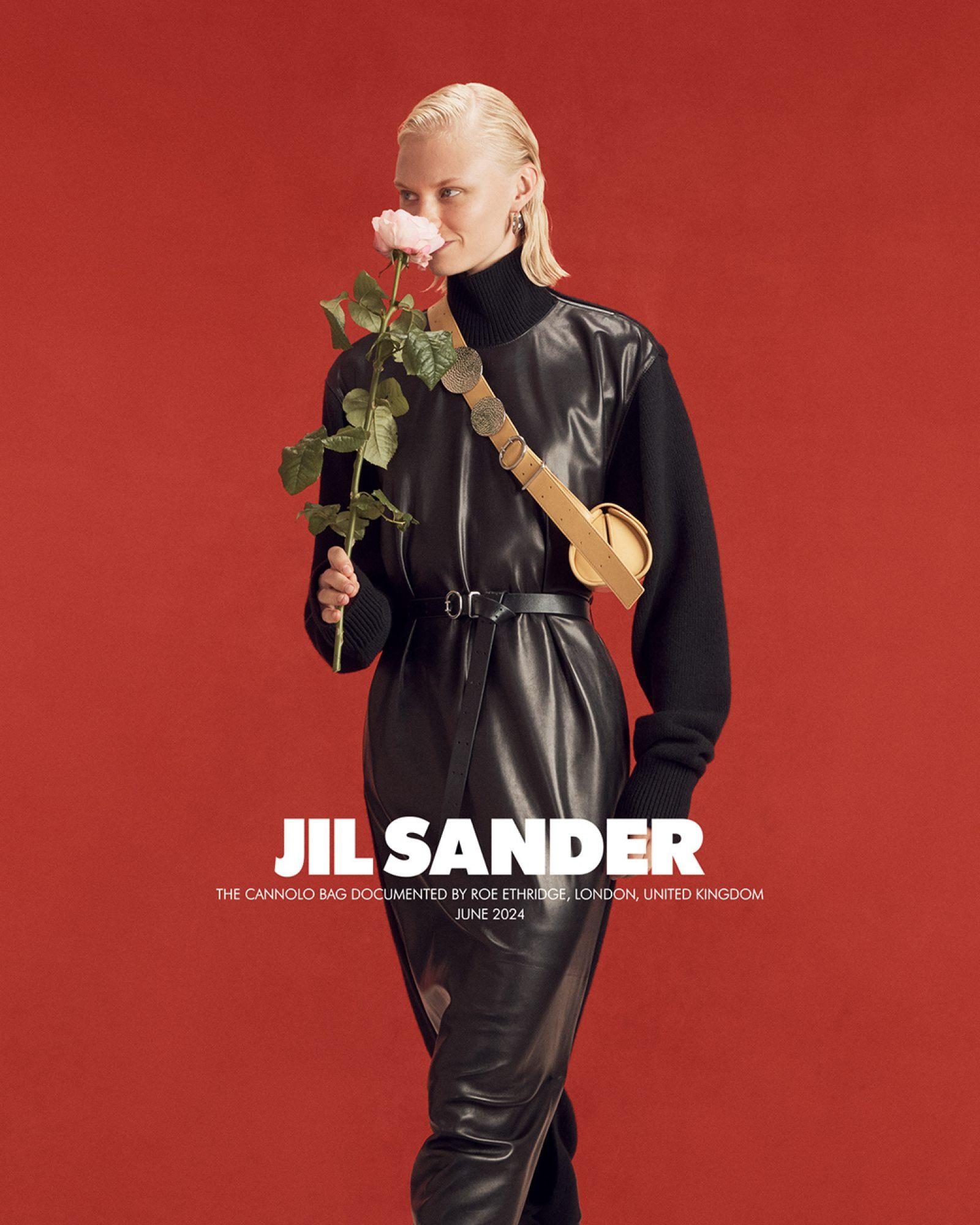 Fashion shot: adult woman poses with a rose, wearing a black leather dress and carrying a tan leather handbag against a red backdrop.
