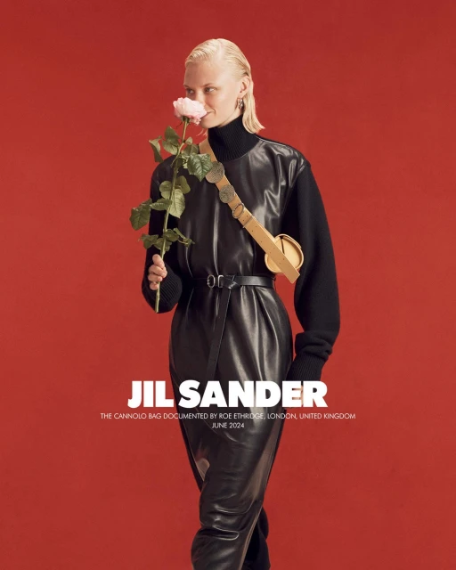 Fashion shot: adult woman poses with a rose, wearing a black leather dress and carrying a tan leather handbag against a red backdrop.