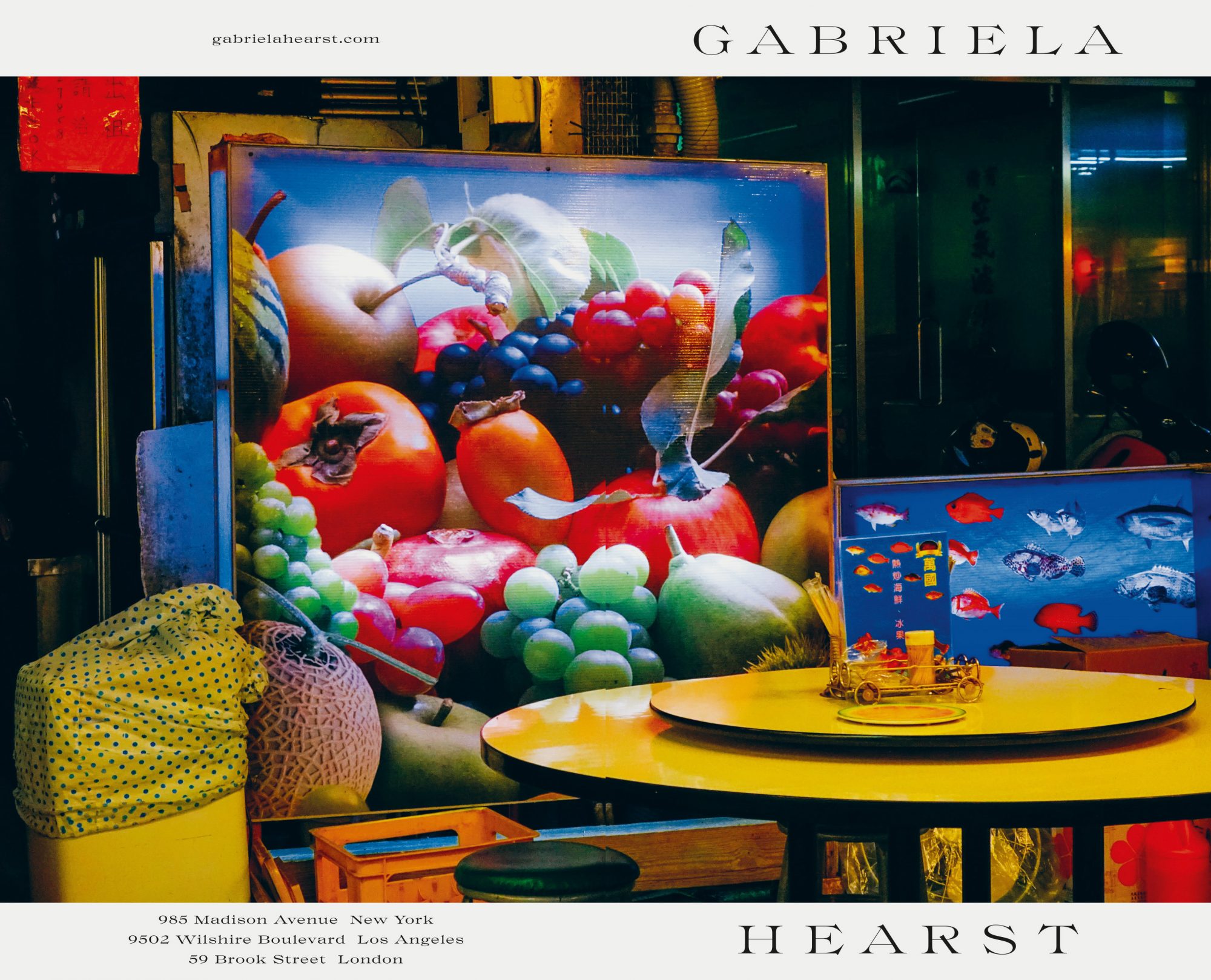 Still life of fruit and fish art displayed next to a yellow table. Shot indoors, the composition features vibrant colors and textures.