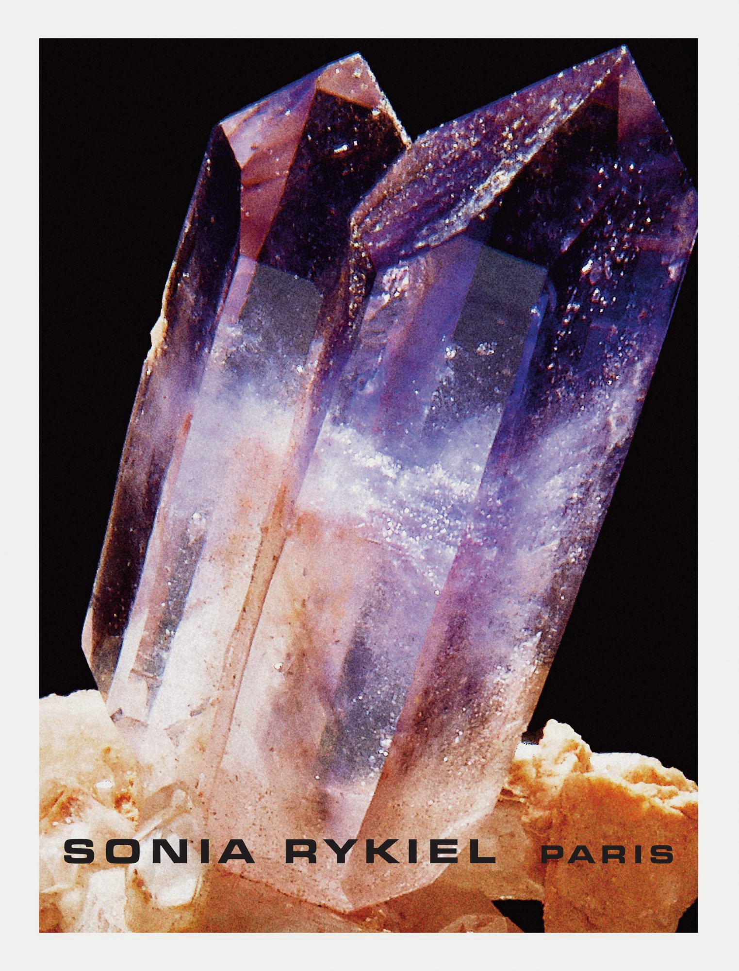Close-up of two amethyst crystals with purple and clear hues, resting on a tan mineral base against a black background, with Sonia Rykiel logo.