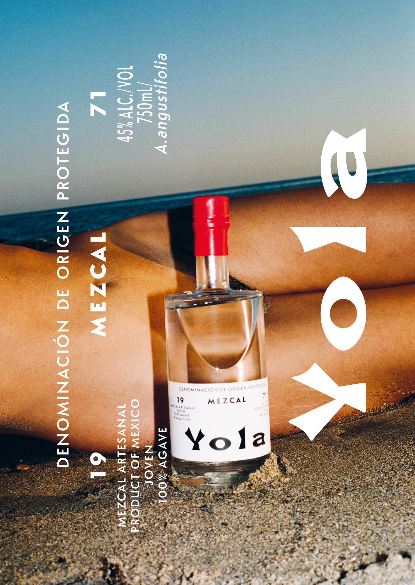 Bottle of Yola mezcal sits on sand in front of a woman lying down by the ocean on a sunny day in an advertising shot.