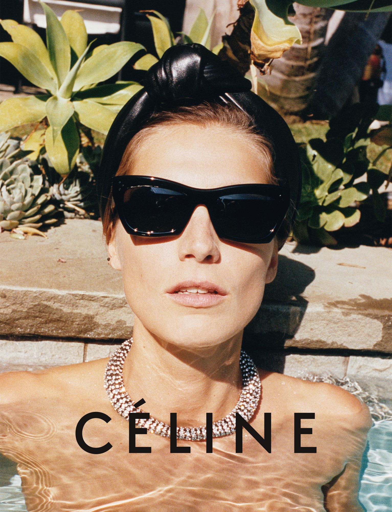 Fashion shot of a woman in a pool wearing sunglasses and a headband, with the Céline logo visible. The background features green plants.