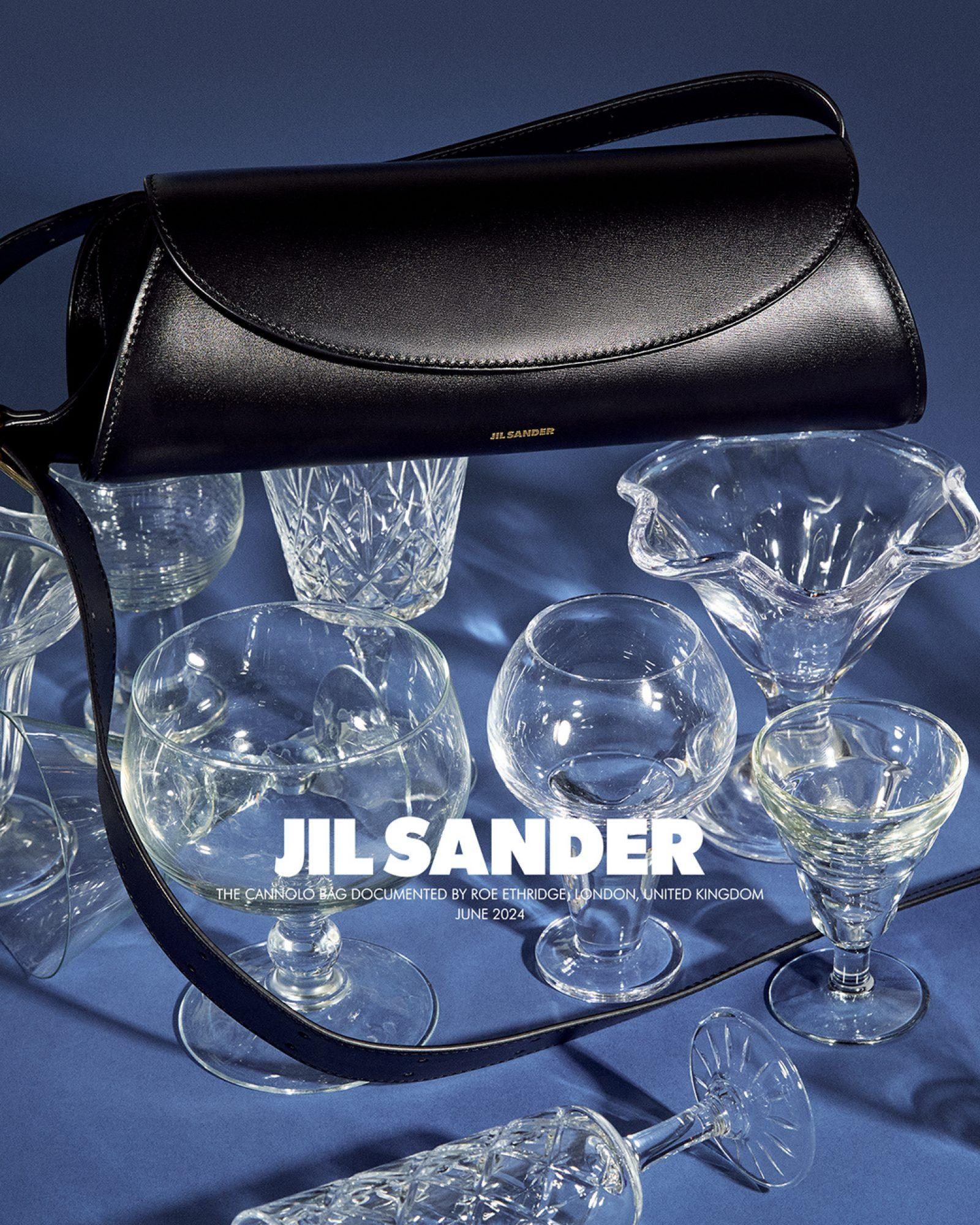 Black Jil Sander handbag on a draped blue fabric, surrounded by an assortment of crystal glassware in a studio setting.